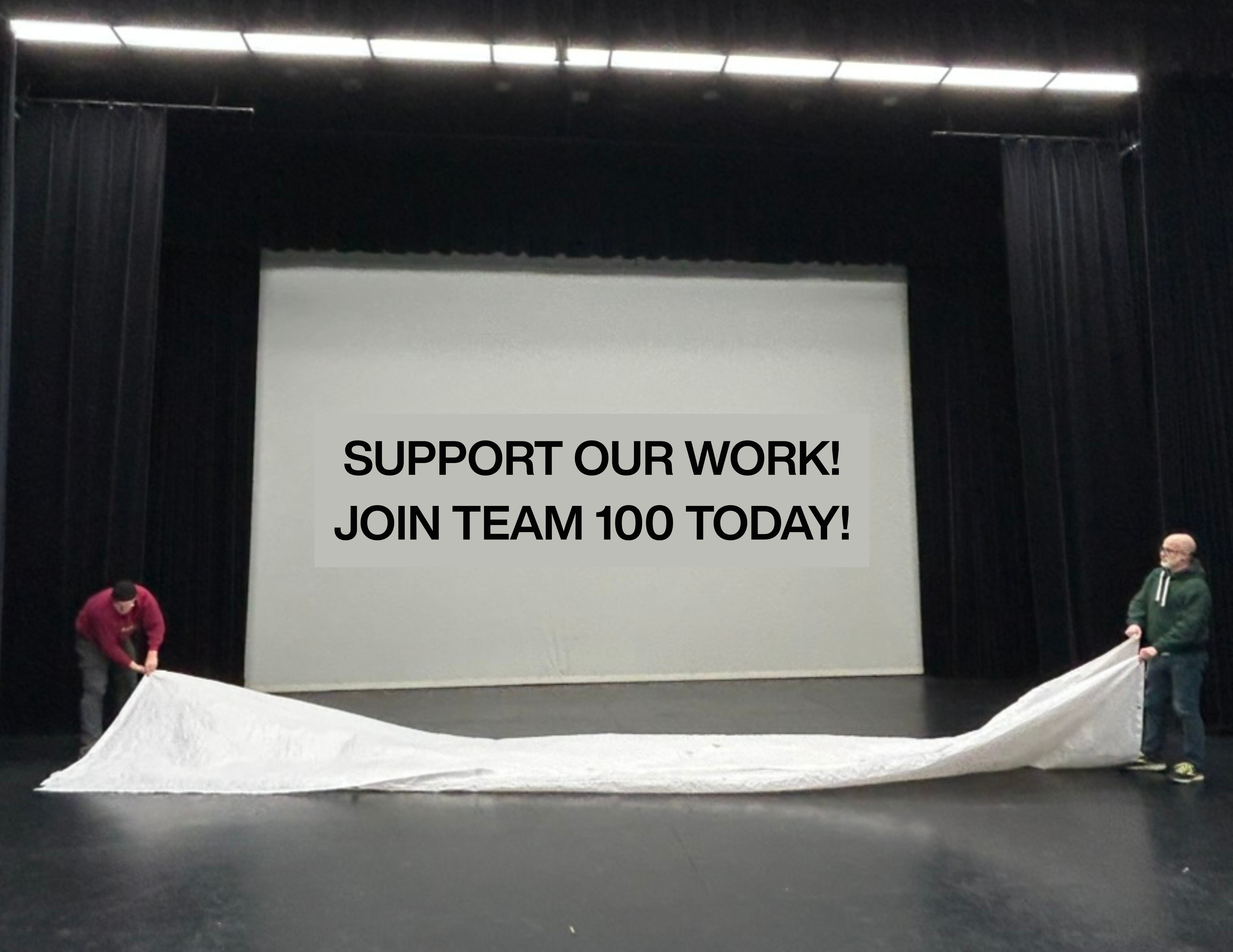 A large white projection screen at the back of a stage. The words "Support Our Work! Join Team 100 Today!" are on it. In the foreground, two men fold a giant white tarp.