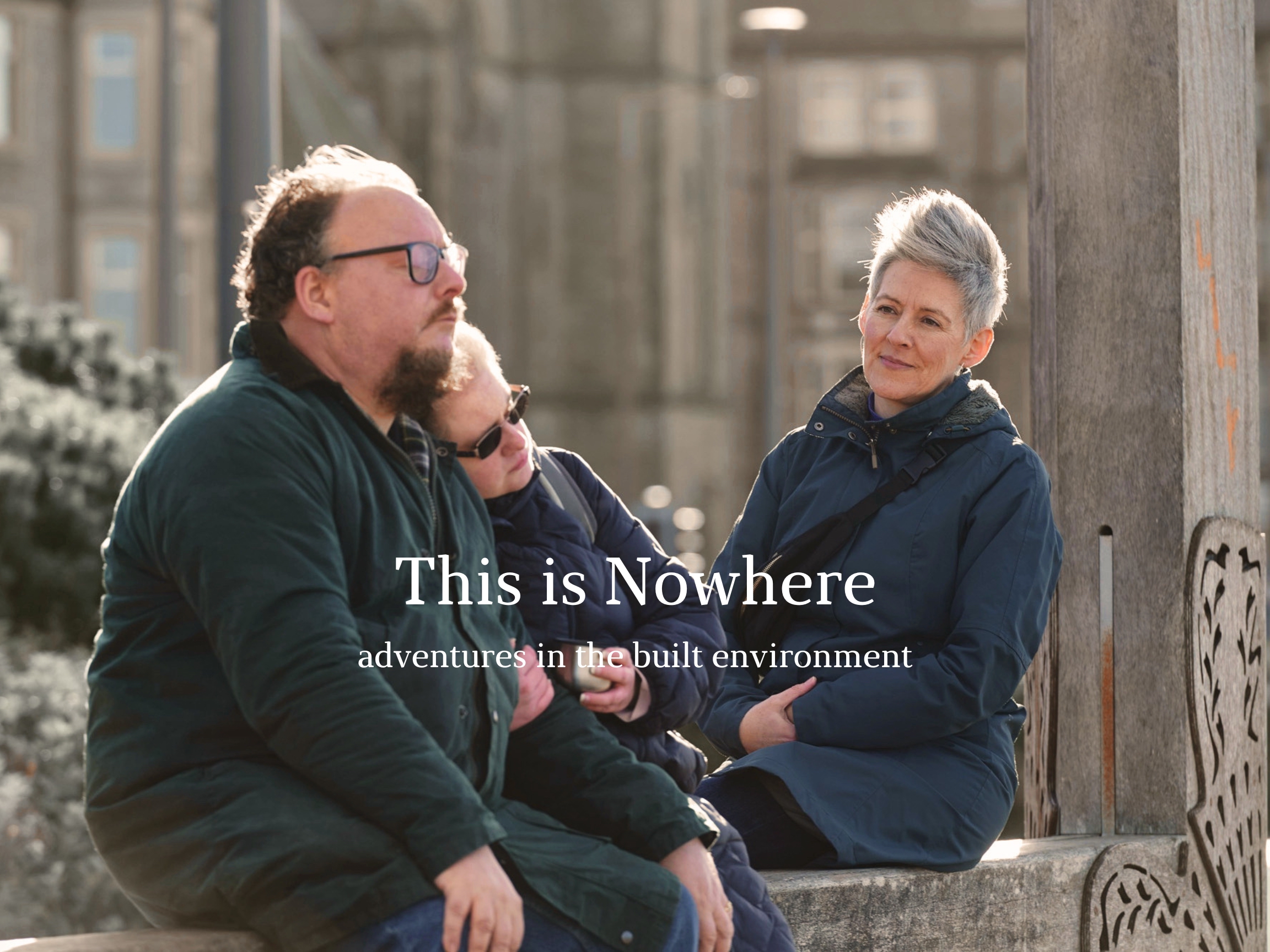 A middle-aged woman leans her head on the shoulder of a middle-aged man, outdoors in a public 'picture frame' installation. They are being watched by a woman sitting on next to them.